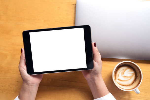Widok z góry makieta kobiecych dłoni trzymających czarną tabletkę z białym, czystym ekranem i laptopem, filiżanka kawy na drewnianym tle stołu - Zdjęcie, obraz