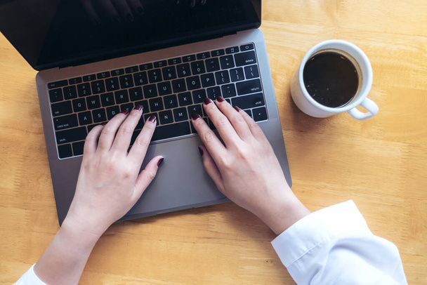 Top view mockup image of woman's hands using and typing on laptop with blank white desktop screen and coffee cup on wooden table in cafe - Photo, image