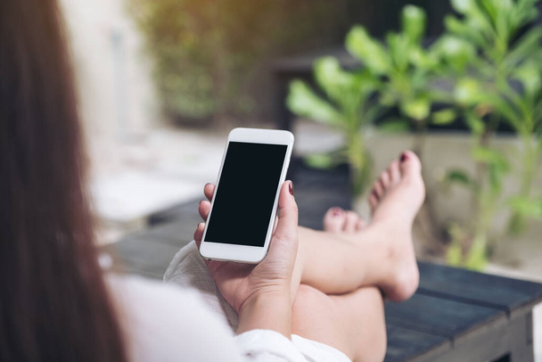 Imagem improvisada de uma mulher sentada e segurando o telefone celular branco com tela de desktop preta em branco, sentindo-se relaxada com areia e fundo de praia
 - Foto, Imagem