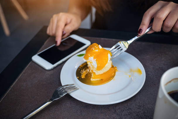 Closeup image of a woman using smartphone while cutting and eating an orange cake with fork on white plate on wooden table in cafe - Foto, Imagem