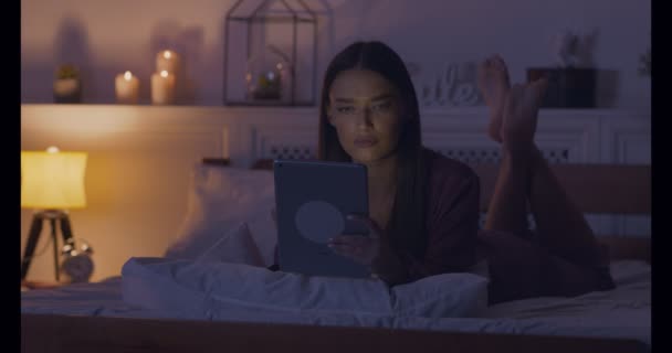 Serious woman reading news on digital tablet, lying on bed - Materiał filmowy, wideo