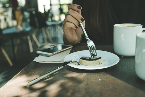Close-up afbeelding van een vrouw snijden een koekje met vork op witte plaat op houten tafel in cafe - Foto, afbeelding