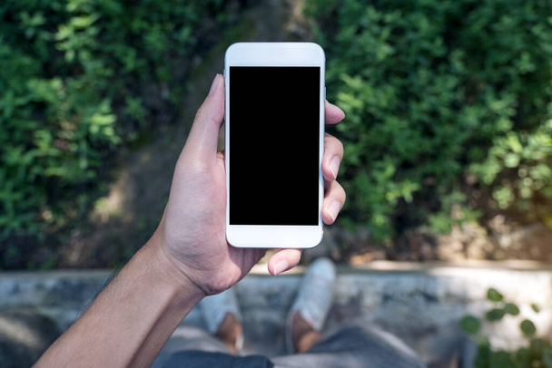 Attrappe eines Mannes, der ein weißes Mobiltelefon mit leerem schwarzen Bildschirm hält, während er auf Betonpolierboden mit grünem Laub und Naturhintergrund steht - Foto, Bild