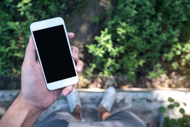 Mockup obraz muže ruka drží bílý mobilní telefon s prázdnou černou obrazovkou, zatímco stojí na betonové lešticí podlahy se zelenou dovolenou a přírodou pozadí - Fotografie, Obrázek