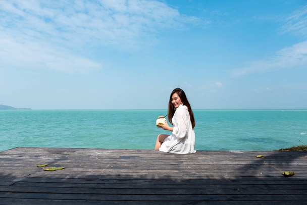 Una hermosa mujer asiática en vestido blanco sentada en la terraza con el mar y el cielo azul con sensación relajada
 - Foto, Imagen
