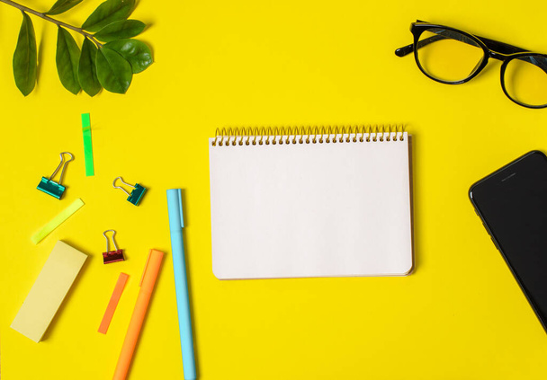 Желтый фон, белая тетрадь для записей, телефон, компьютерные очки, растения, ручки.
 - Фото, изображение