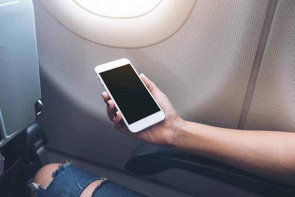 Bir uçağın penceresinin yanında boş siyah ekranlı beyaz akıllı bir telefon tutan kadınların görüntüsü.  - Fotoğraf, Görsel