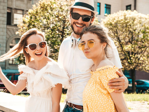 Group of young three stylish friends posing in the street. Fashion man and two cute girls dressed in casual summer clothes. Smiling models having fun in sunglasses.Cheerful women and guy outdoors - Foto, Imagen