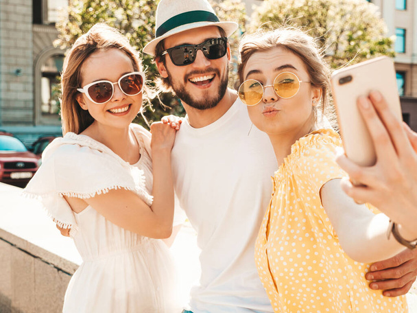 Group of young three stylish friends in the street.Man and two cute girls dressed in casual summer clothes.Smiling models having fun in sunglasses.Women and guy making photo selfie on smartphone - Zdjęcie, obraz