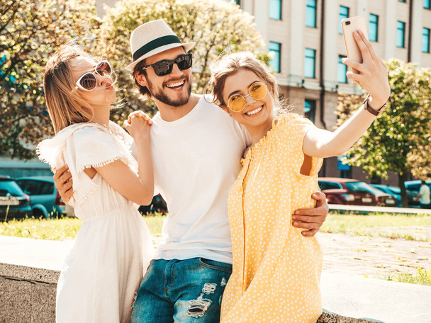 Group of young three stylish friends in the street.Man and two cute girls dressed in casual summer clothes.Smiling models having fun in sunglasses.Women and guy making photo selfie on smartphone - Photo, image