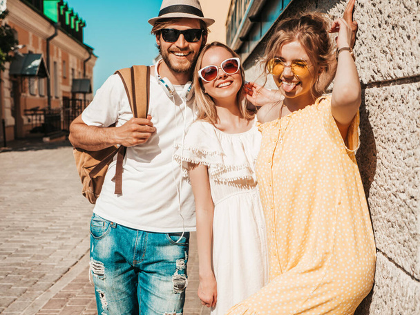 Group of young three stylish friends posing in the street. Fashion man and two cute girls dressed in casual summer clothes. Smiling models having fun in sunglasses.Cheerful women and guy outdoors - 写真・画像