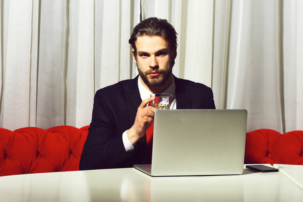 bearded man, businessman holding glass of whiskey works at laptop - 写真・画像