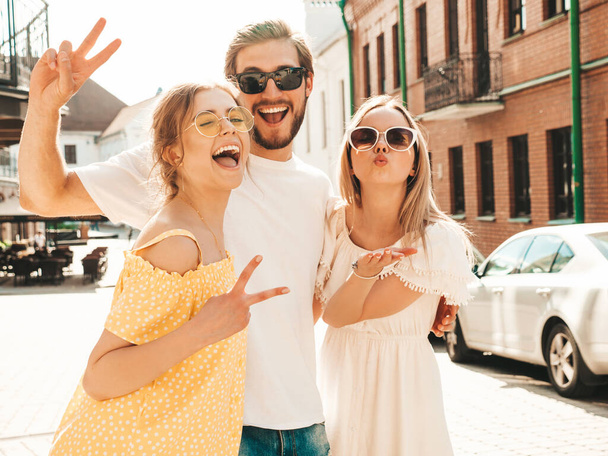 Group of young three stylish friends posing in the street. Fashion man and two cute girls dressed in casual summer clothes. Smiling models having fun in sunglasses.Cheerful women and guy going crazy - Photo, Image
