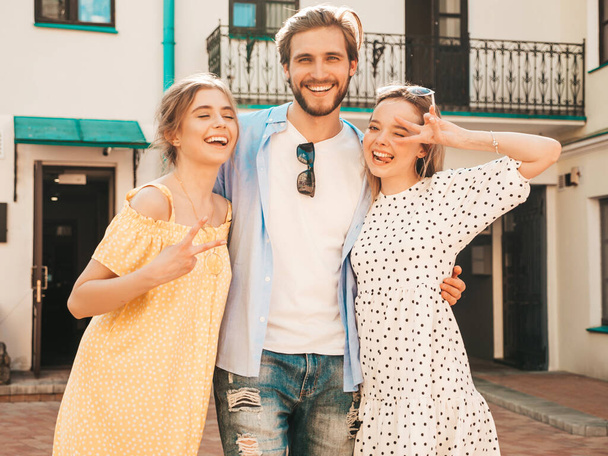 Group of young three stylish friends posing in the street. Fashion man and two cute girls dressed in casual summer clothes. Smiling models having fun in sunglasses.Cheerful women and guy going crazy - Foto, Imagen