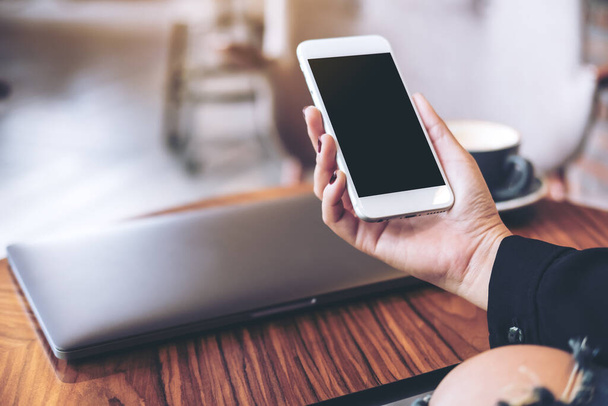 Imagem Mockup da mão de uma mulher segurando telefone celular branco com tela de desktop preta em branco e laptop na mesa de madeira no café
 - Foto, Imagem
