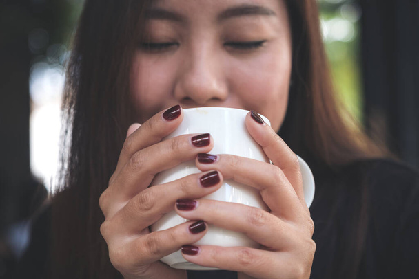 Imagen de cerca de una hermosa mujer asiática cerrando los ojos y sosteniendo una taza blanca de café caliente con sentirse feliz en la cafetería
 - Foto, imagen