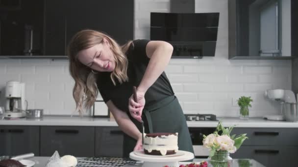 Professional chef is cooking cake. Young attractive housewife uses metal spatula and rotating table to aligns white cream on chocolate cake - Footage, Video