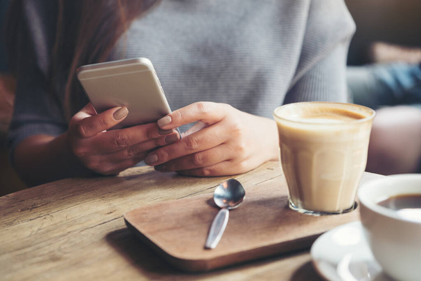 Imagen de cerca de una mujer sosteniendo y usando un teléfono inteligente con tazas de café en la mesa de madera en la cafetería
 - Foto, Imagen