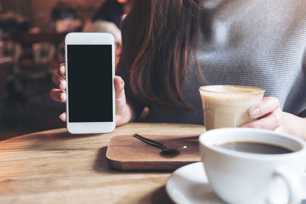 Immagine mockup di una donna che tiene e mostra il telefono cellulare bianco con schermo desktop nero vuoto con tazze da caffè su tavolo in legno vintage - Foto, immagini