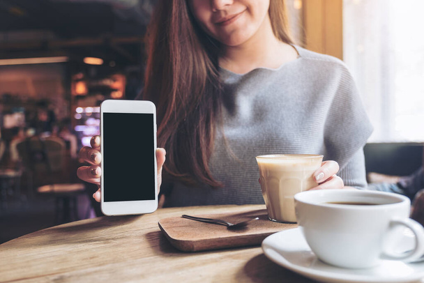 ヴィンテージ木製テーブルの上にコーヒーカップと空白の黒いデスクトップ画面と白い携帯電話を保持し、示す美しい女性のモックアップイメージ - 写真・画像