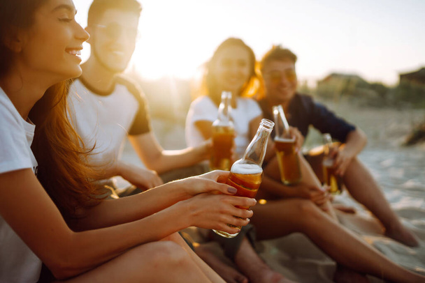 Young people sitting together at beach, drinking beer and having a party. Group of friends cheers with beers at the beach during summer vacation. - Foto, Imagen