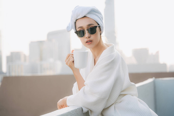 Beyaz bornozlu, güneş gözlüğü takan genç bir kadın sabahları apartman dairesinde kahve içmenin keyfini çıkarıyor. Şehir arkaplanı. - Fotoğraf, Görsel