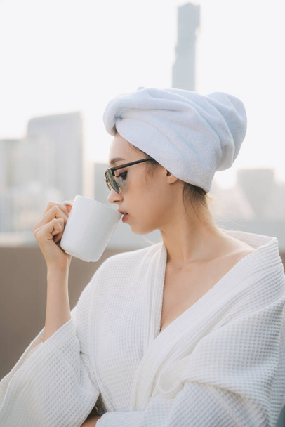 Oldalnézet - Fiatal nő fehér fürdőköpenyben napszemüvegben forró kávét iszik otthon. - Fotó, kép