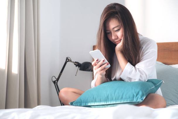 Portret pięknej Azjatki siedzącej na łóżku, korzystającej z telefonu komórkowego i patrzącej na niego po przebudzeniu z poczuciem szczęścia - Zdjęcie, obraz