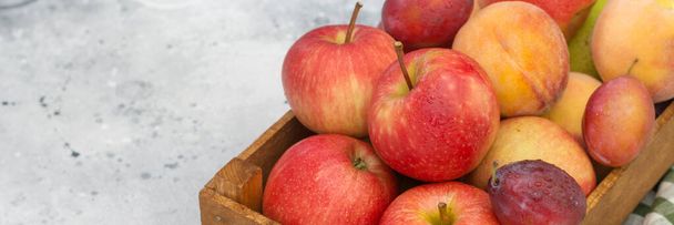 Various fruits in a wooden box on the light gray kitchen table. Apples, plums and peaches in a brown wooden box. Ripe raw fruit close-up. Banner with space for text - Foto, imagen
