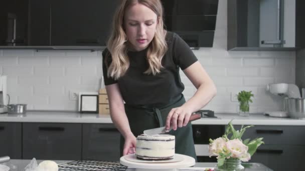 プロのシェフがケーキを作っています。若い魅力的な主婦はチョコレートケーキの上に白いクリームを整列させるために金属へらと回転テーブルを使用します - 映像、動画