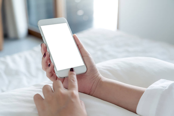 Mockup image of a woman's hand holding and touching white mobile phone with blank desktop screen sitting in the white bed - Foto, immagini