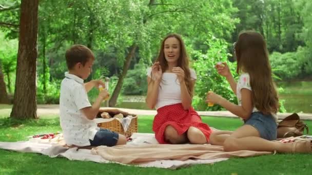 Girl and boy blowing bubbles in summer park. Smiling woman relaxing with kids - Materiał filmowy, wideo