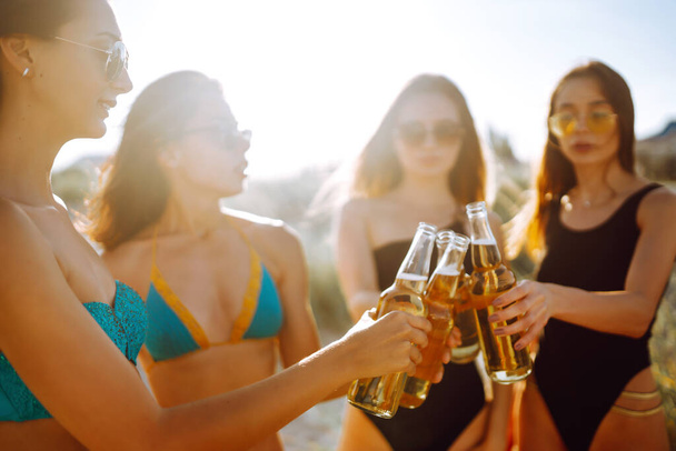Four beautiful girls cheers and drink beers on the beach, enjoying vacation. Vacation time laughing and smiling. - Foto, Imagen