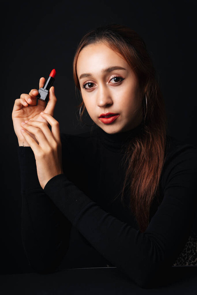 Close up portrait of pretty beautiful young woman wearing black sweater isolate over dark background. Beauty in the dark. Woman with cosmetic red lipstick on hand. - 写真・画像