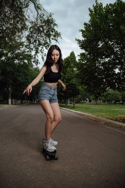 Young girl riding a skateboard in the city.Young woman in a black tank top and blue shorts rides on a skateboard in the city.Portrait young woman riding skateboard outdoors lifestyle - Fotografie, Obrázek