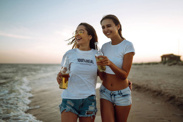Νεαρά κορίτσια περπατάνε στην παραλία και πίνουν μπύρες το ηλιοβασίλεμα, κατά τη διάρκεια των καλοκαιρινών διακοπών. Διακοπές, να χαλαρώσετε και τον τρόπο ζωής έννοια. - Φωτογραφία, εικόνα