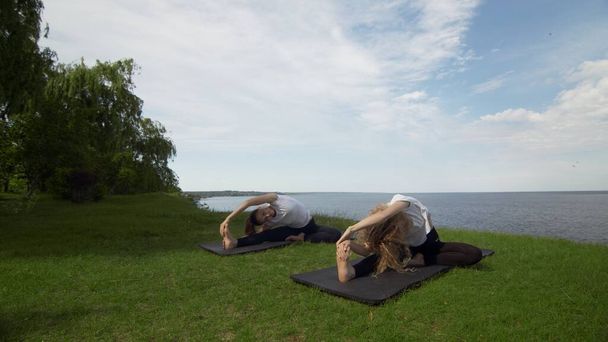 Két nő meditál a szabadban ül a part szélén a tenger közelében, fél lótusz összecsukott ujjakkal mudra gesztus jóga gyakorlat reggel. Két nő gyakorolja a Forgatott Fej a Térdig pózt - Fotó, kép