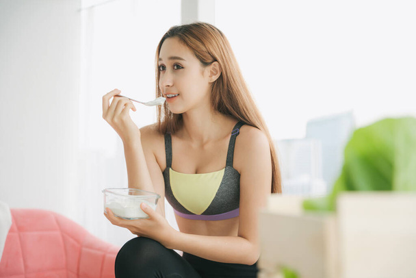 Beautiful woman in sportwear with natural make up isolated over white background. Healthy woman in sportbra enjoy eating yogurt with spoon from glass bowl on a couch. Quarantine at home. - Photo, Image