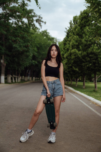 Young woman posing on street with skateboard in hands.young girl in a black tank top and blue shorts posing with a skateboard.Concept of youth recreation, lifestyle. Skateboarder in the city. - 写真・画像