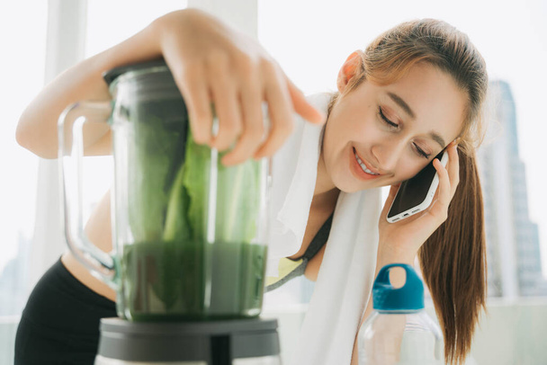 Hermosa mujer sana haciendo jugo de verduras saludables con mezcladora y hablando por teléfono con un amigo dentro de la cocina
. - Foto, Imagen
