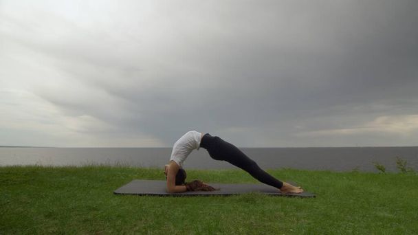 Młoda, sprawna kobieta ćwiczy jogę na wybrzeżu w pobliżu jeziora lub morza. Kobieta robi odwrócony personel Dvi Pada Viparita Dandasana pozować - Zdjęcie, obraz
