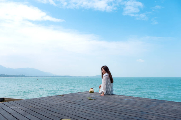 A beautiful asian woman on white dress sitting at the terrace with sea and blue sky background with feeling relaxed - Photo, Image