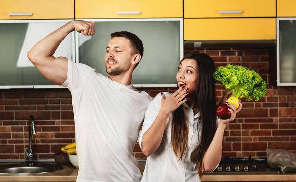 Amigos felices preparando comida para la cena, posando en la cocina sosteniendo verduras y frutas. El hombre muestra bíceps en su brazo. Promover una alimentación saludable
 - Foto, imagen