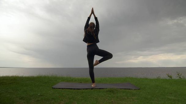 Mladá žena v černé mikině cvičí jógu na pobřeží u jezera nebo u moře. Žena dělá strom póza Vrksasana Vriksasana Pose - Fotografie, Obrázek