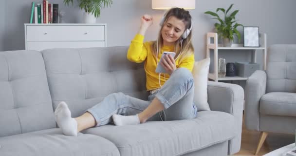 Woman enjoying music from her phone with white headphones and dancing in the sofa, having fun in a beautiful minimalistic living room. The concept of modern life, relaxation, lifestyle. - Video