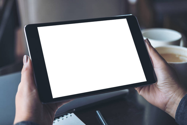 Mockup afbeelding van handen met zwarte tablet pc met blanco witte desktop scherm met koffiebekers op tafel in een modern cafe - Foto, afbeelding