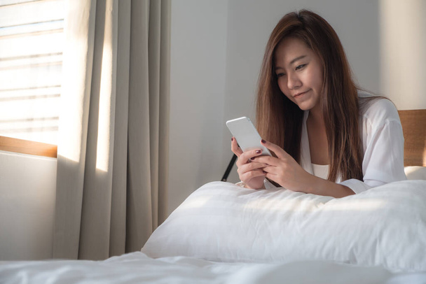 Портретное изображение красивой азиатки, сидящей на кровати и смотрящей на мобильный телефон после пробуждения с чувством счастья
 - Фото, изображение