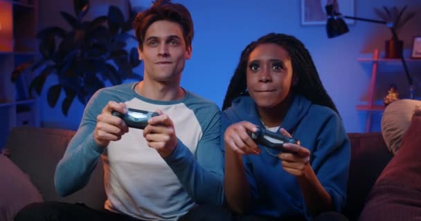 Happy boyfriend and girlfriend playing video games and holding joysticks at home. Millennial couple enjoing game while sitting on sofa and spending free time. Concept of relationship. - Materiał filmowy, wideo