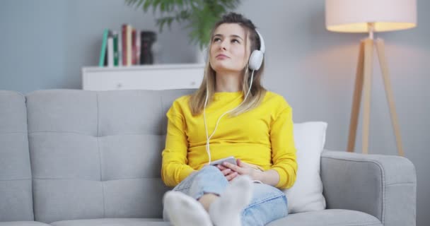 Cheerful, playful girl listening music at phone using headphones, moving head in rhytm. Woman having fun in beautiful minimalistic living room. Concept of self-development, relax, lifestyle. - Πλάνα, βίντεο