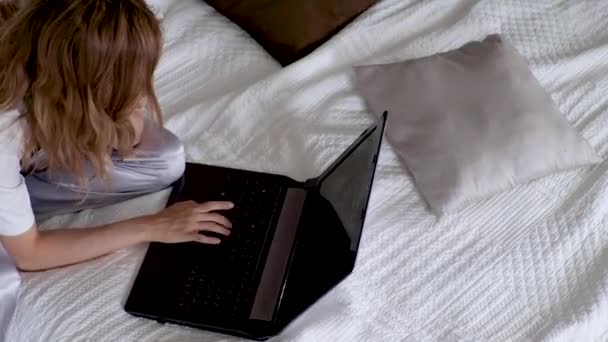 A young woman works remotely. A girl in pajamas and a t-shirt is sitting on the bed - Video